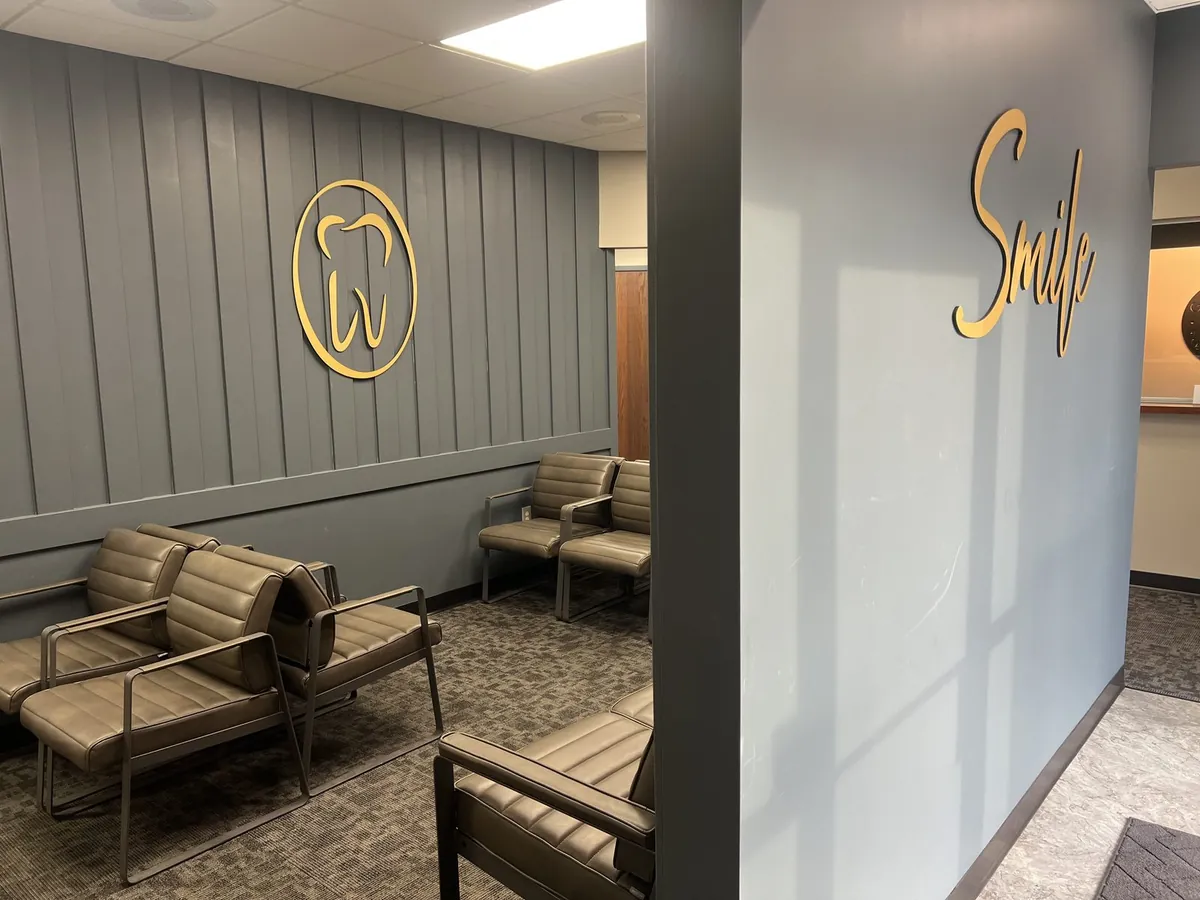 The AAA Court Family Dental waiting room, modern grey leather seating, gold tooth logo on a charcoal accent wall, and a gold 'Smile' script greeting patients